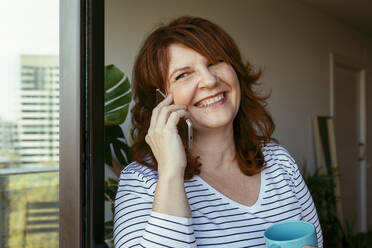 Lächelnde Frau, die mit ihrem Handy telefoniert, während sie zu Hause am Fenster steht - VABF03564