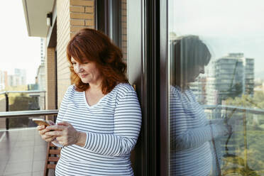 Woman using mobile phone while standing in balcony at home - VABF03565