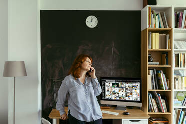 Frau, die am Schreibtisch stehend zu Hause mit ihrem Handy telefoniert - VABF03567