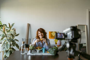 Smiling woman showing fresh fruits while making vlog on camera at home - VABF03574