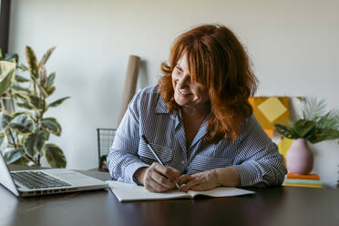 Lächelnde Frau schreibt ein Buch, während sie zu Hause am Tisch sitzt - VABF03579