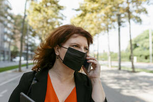 Woman with face mask talking on mobile phone while standing on road in city - VABF03581