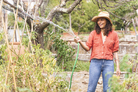 Smiling beautiful mature woman watering plants from hose at garden stock photo