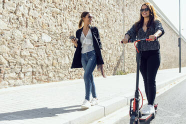 Woman walking by friend riding push scooter on road in city - JSRF01150