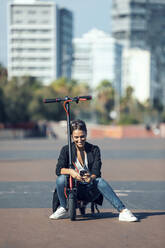 Young woman using mobile phone while sitting on push scooter in city - JSRF01156