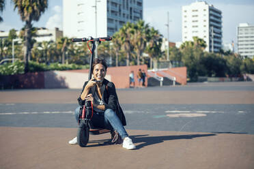 Smiling woman sitting on push scooter in city - JSRF01157