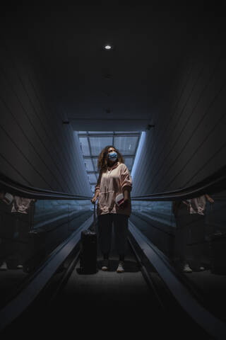 Young woman wearing protective face mask standing on escalator during covid-19 stock photo