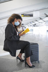 Young woman wearing face mask reading book while sitting at airport - SNF00599