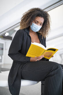 Frau mit lockigem Haar liest ein Buch und trägt eine Gesichtsmaske, während sie am Flughafen sitzt - SNF00600