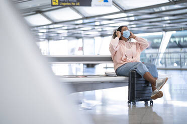 Young woman listening to music while wearing protective face mask sitting at airport - SNF00609