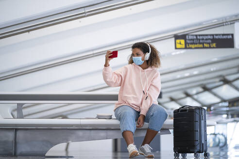 Junge Frau, die ein Selfie macht, während sie über Kopfhörer am Flughafen Musik hört - SNF00611