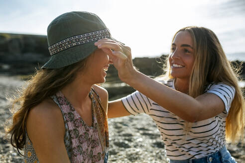 Lächelnde junge Frau, die den Hut ihrer Schwester zurechtrückt, während sie ihr Wochenende am Strand genießt - MGOF04483