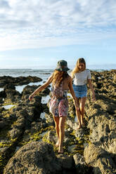 Female friends walking on rocks at beach against sky - MGOF04491