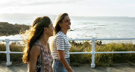 Smiling young women walking on footpath on sunny day - MGOF04498