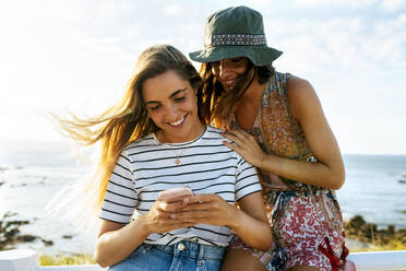 Lächelnde junge Freundinnen benutzen ihr Smartphone am Meer während eines sonnigen Tages - MGOF04502