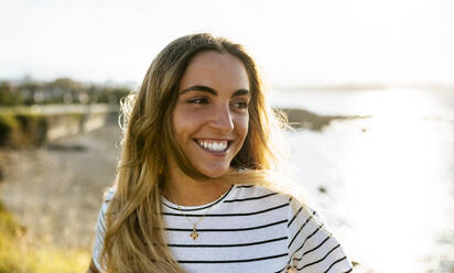 Glückliche junge Frau schaut weg, während sie ein Wochenende am Strand bei Sonnenschein verbringt - MGOF04506