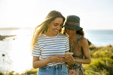Smiling young woman showing smart phone to friend while enjoying weekend at beach - MGOF04507
