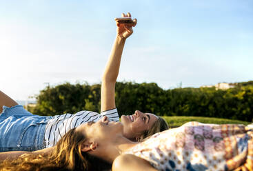 Lächelnde junge Frau, die ein Selfie mit einer Freundin macht, während sie sich auf einem Feld an einem sonnigen Tag entspannt - MGOF04514