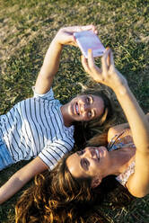 Glückliche junge Frauen, die ein Selfie machen, während sie sich auf einem Feld an einem sonnigen Tag entspannen - MGOF04515