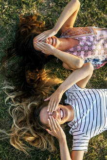 Fröhliche junge Frauen bedecken ihre Augen mit den Händen, während sie auf einem Feld liegen - MGOF04519