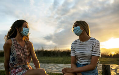 Frauen mit Gesichtsschutz, die während des Sonnenuntergangs gegen den Himmel sitzen und soziale Distanz wahren - MGOF04522