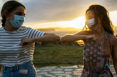 Young women in protective face masks elbow bumping against sky during COVID-19 - MGOF04524