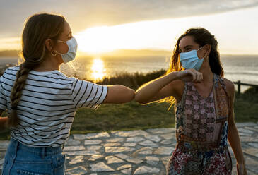 Women in protective face masks elbow bumping against sky during COVID-19 outbreak - MGOF04525