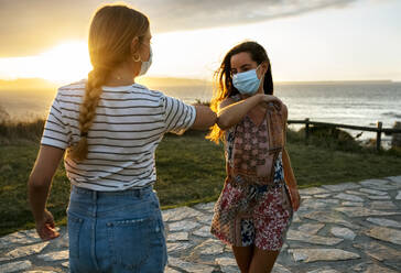 Female friends in protective face masks elbow bumping on footpath during COVID-19 outbreak - MGOF04526