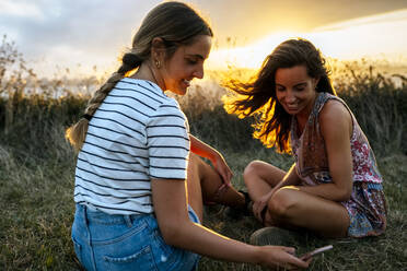 Smiling young woman showing smart phone to female friend while sitting on field during sunset - MGOF04528