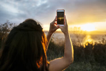 Junge Frau fotografiert Sonnenuntergang Himmel durch Smartphone - MGOF04531