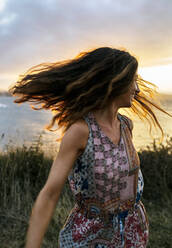 Fröhliche junge Frau, die bei Sonnenuntergang ihr Haar gegen den Himmel wirft - MGOF04533