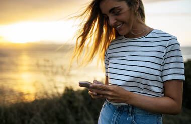 Lächelnde junge Frau mit Smartphone am Strand gegen den Himmel bei Sonnenuntergang - MGOF04534