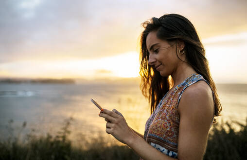 Lächelnde schöne Frau mit Smartphone am Strand gegen den Himmel bei Sonnenuntergang - MGOF04535