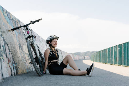 Cheerful young female in activewear and helmet sitting on asphalt path near bicycle and stretching legs while preparing for trip in summer day - ADSF16148