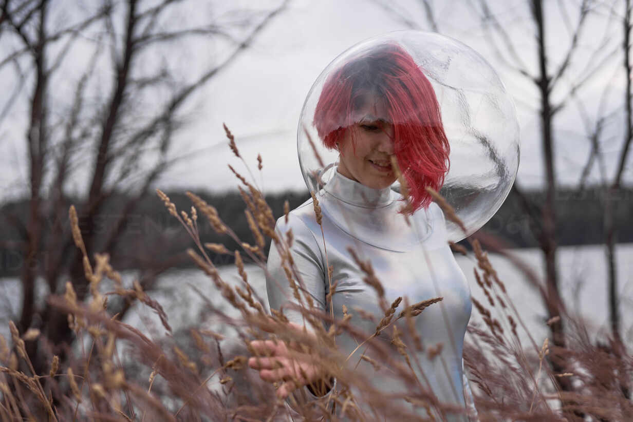 Futuristische junge rothaarige Frau in silbernem Raumanzug und Glashelm  geht in der Nähe eines überfluteten Flusses spazieren, lizenzfreies  Stockfoto