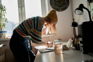 Seitenansicht einer begeisterten Künstlerin in modischer Kleidung, die im Atelier ein Kunstwerk schafft, während sie mit Wasserfarben auf Papier malt - ADSF16162