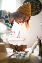 Seitenansicht einer begeisterten Künstlerin in modischer Kleidung, die im Atelier ein Kunstwerk schafft, während sie mit Wasserfarben auf Papier malt - ADSF16164