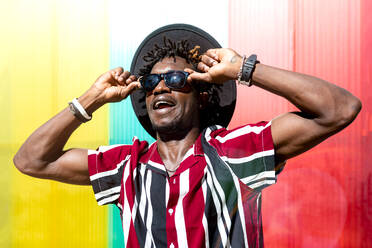 Young African American male in striped shirt and trendy sunglasses and hat looking at camera while standing against multicolored wall - ADSF16179