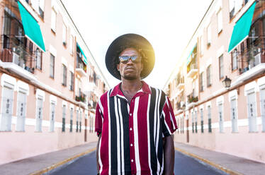 Low angle of confident young African American male in stylish stripped shirt and sunglasses with hat looking at camera while standing against blurred urban street - ADSF16181