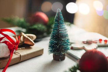 Kleiner grüner Tannenbaum Spielzeug in der Nähe von Weihnachtsgeschenk in Bastelpapier eingewickelt auf dem Tisch mit Dekorationen gegen unscharfe leuchtende Girlande platziert - ADSF16226