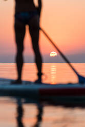 Back view of crop anonymous woman standing on surfboard and rowing with paddle during training on background of amazing sundown sky - ADSF16242