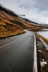 Spektakulärer Blick auf eine nasse, kurvenreiche Straße in Flussnähe und einen Berghang mit trockenem Gras im Winter auf den Färöern - ADSF16254