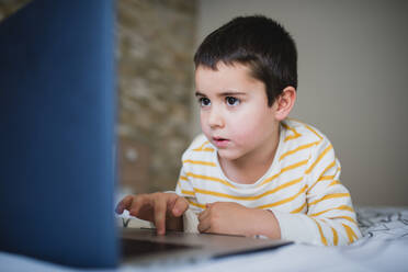 Focused little boy in casual wear browsing laptop while spending free time in bedroom at home - ADSF16280