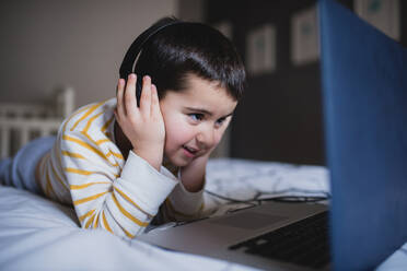 Focused little boy in casual wear browsing laptop while spending free time in bedroom at home - ADSF16282