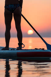 Back view of crop anonymous woman standing on surfboard and rowing with paddle during training on background of amazing sundown sky - ADSF16294