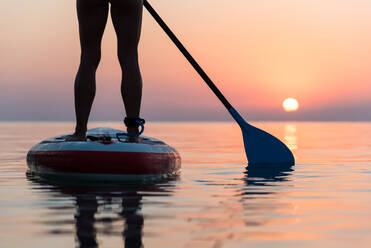 Back view of crop anonymous woman standing on surfboard and rowing with paddle during training on background of amazing sundown sky - ADSF16295