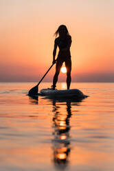 Rückansicht der Silhouette einer nicht erkennbaren weiblichen Surferin, die auf einem Paddelbrett steht und gegen die spektakuläre Sonne im Sonnenuntergang rudert - ADSF16297