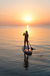 Back view of silhouette of unrecognizable female surfer standing on paddleboard and rowing against spectacular sun in sunset sky - ADSF16299