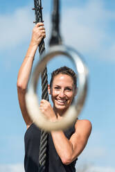Muscular female athlete climbing rope while training on sports ground in city and looking at camera - ADSF16341