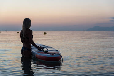 Side view of of unrecognizable female surfer standing near paddleboard and rowing against spectacular sun in sunset sky - ADSF16344